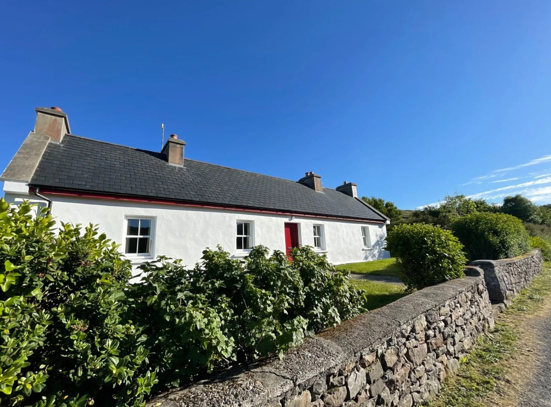 Angled view of Clare Island Farmhouse with whitewashed walls, a red door, garden planting, and a stone wall