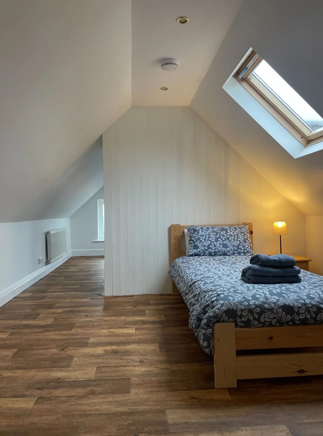 Detail of the Room 4 twin loft room with patterned bedding, bedside lamp, folded towels, and roof window