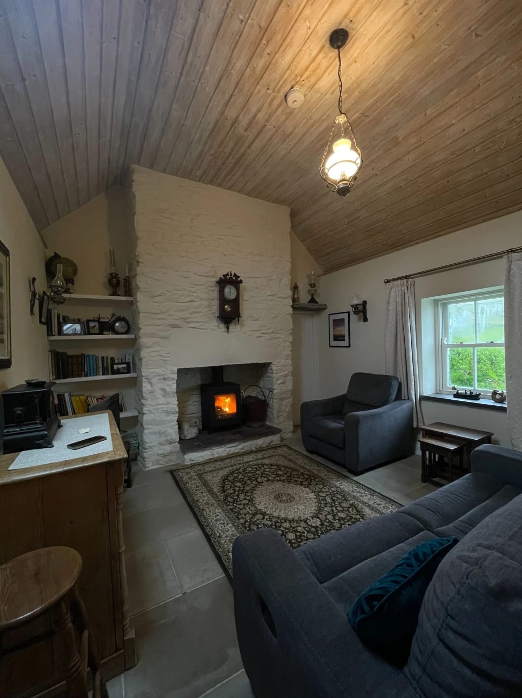 Shared farmhouse sitting room with a stone fireplace, lit stove, armchairs, timber ceiling, and bookshelves
